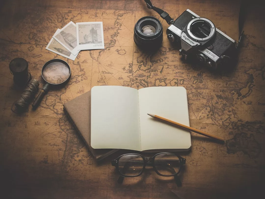 An open notebook with a pencil, eyeglasses, camera, photos, lens, magnifying glass, and twine rest on an old world map—capturing the essence of a wanderer’s lifestyle.