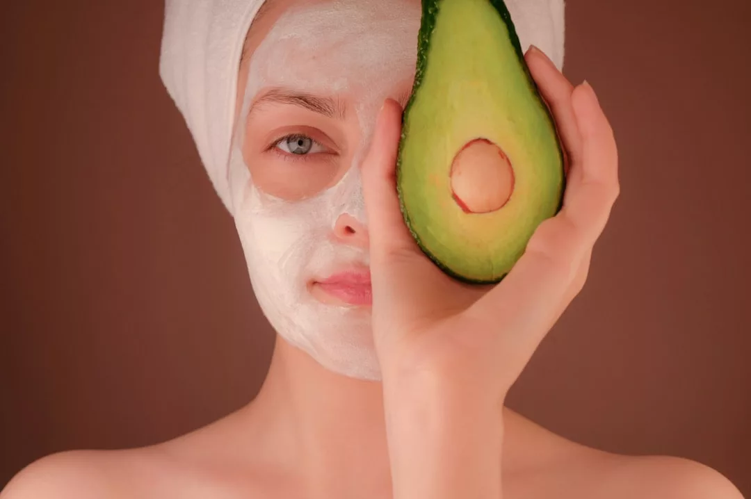 A person with a white facial mask and a towel on their head holds half an avocado in front of one eye, capturing a playful moment of self-care and luxury lifestyle against a plain background.